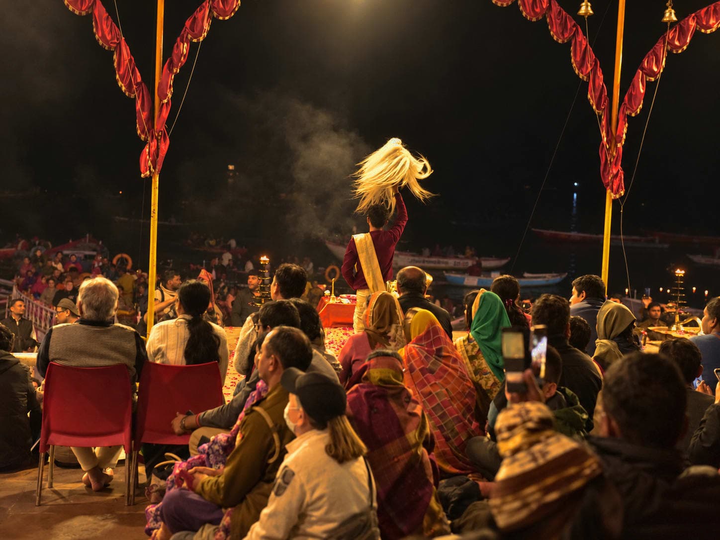 A priest raises a white ceremonial whisk above the seated crowd during evening Ganga aarti in Varanasi, 2019