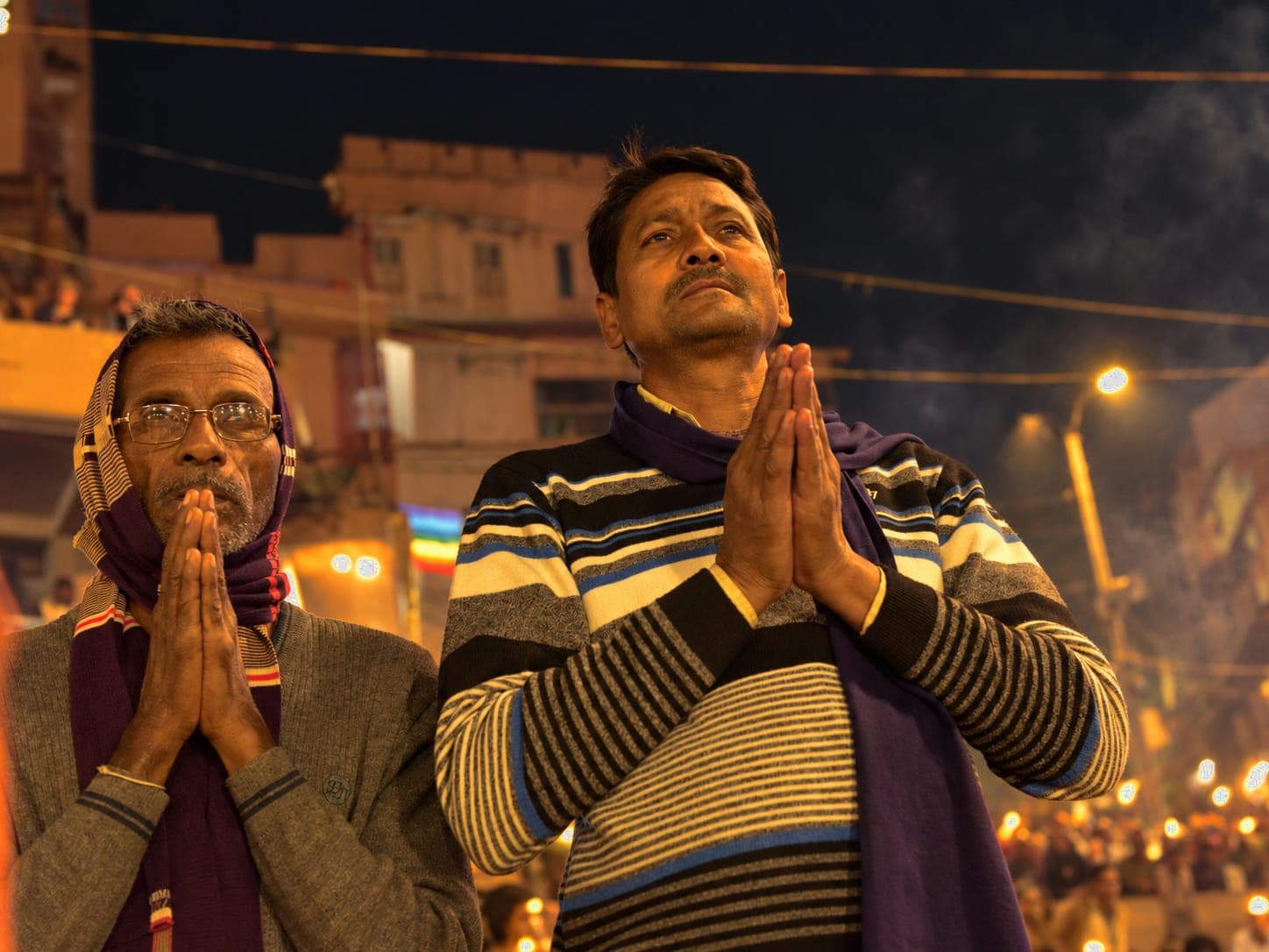 Two men hold their palms together in prayer while looking upward during evening worship in Varanasi, 2019