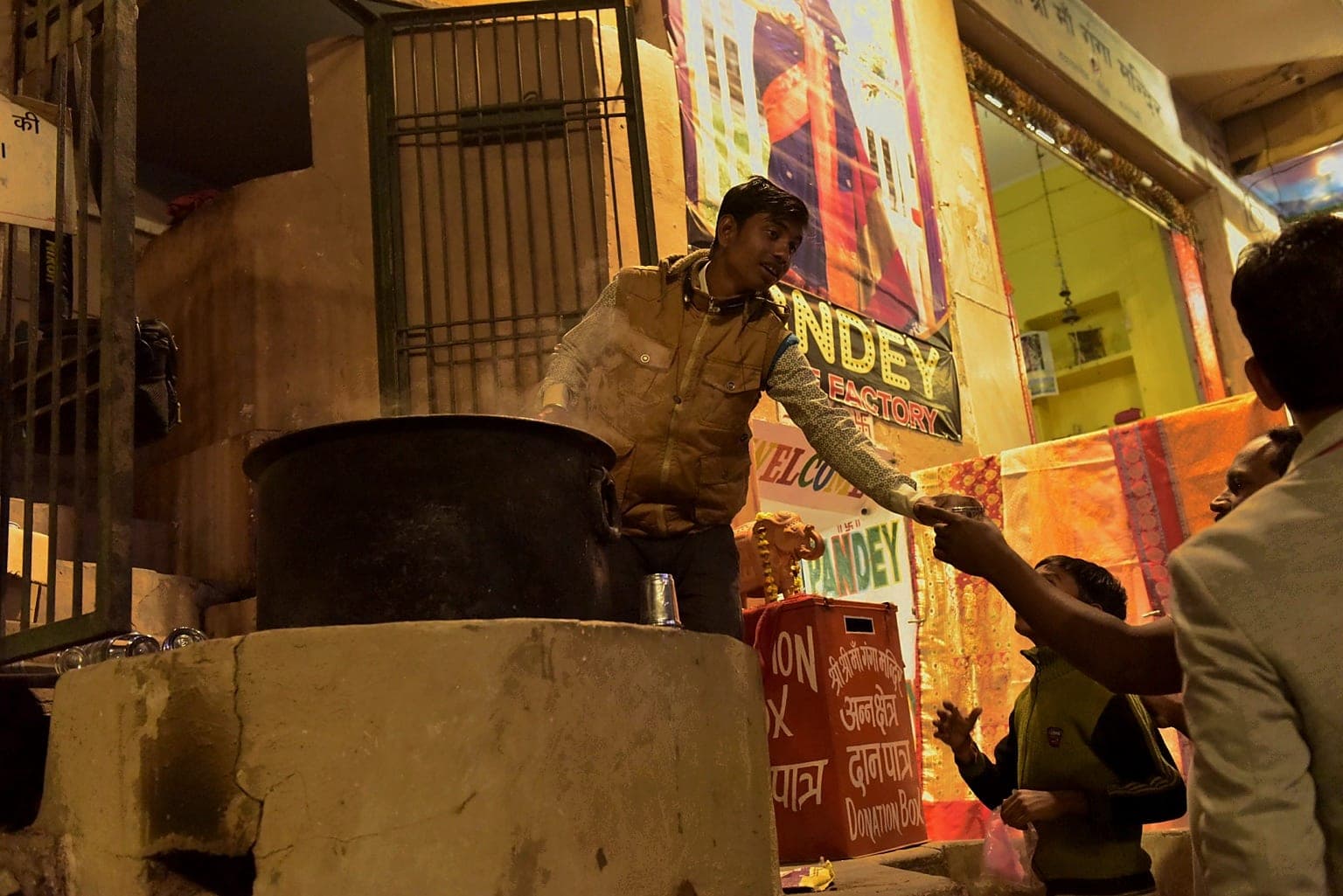 A young man reaches to exchange an item beside a large cooking vessel and a donation box at night in Varanasi, 2019
