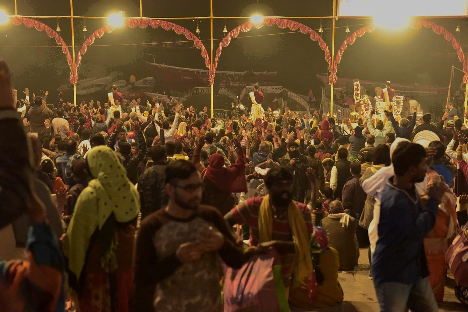 A dense crowd raises hands toward the riverfront ceremony beneath illuminated arches in Varanasi, 2019