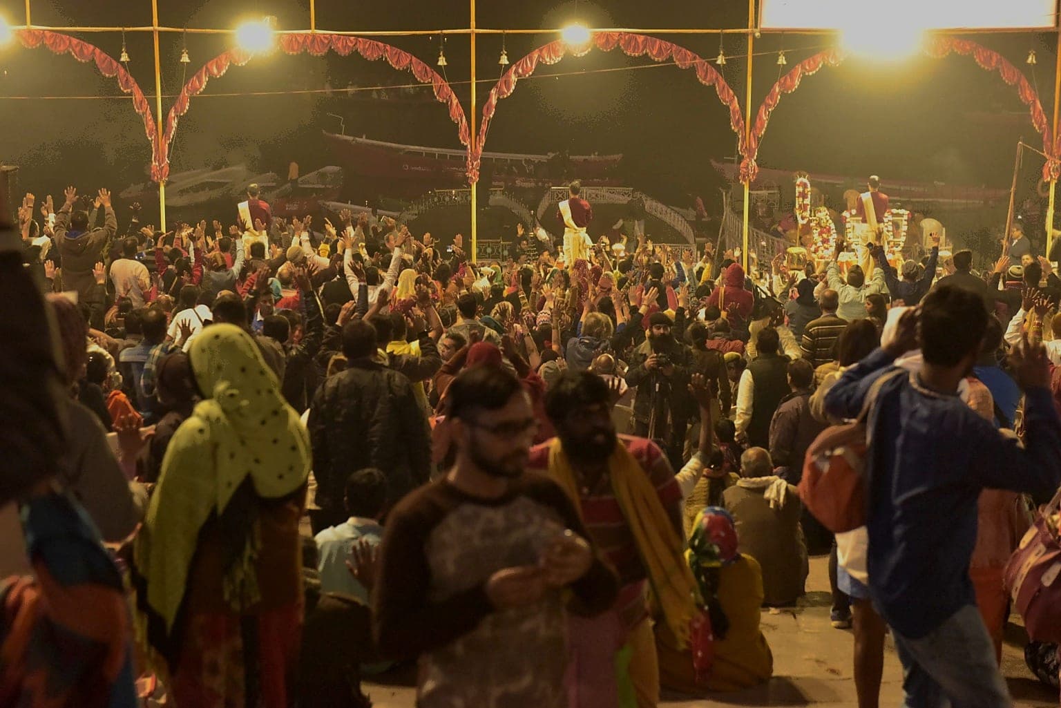 A large night crowd faces the riverfront platforms with many hands raised in Varanasi, 2019