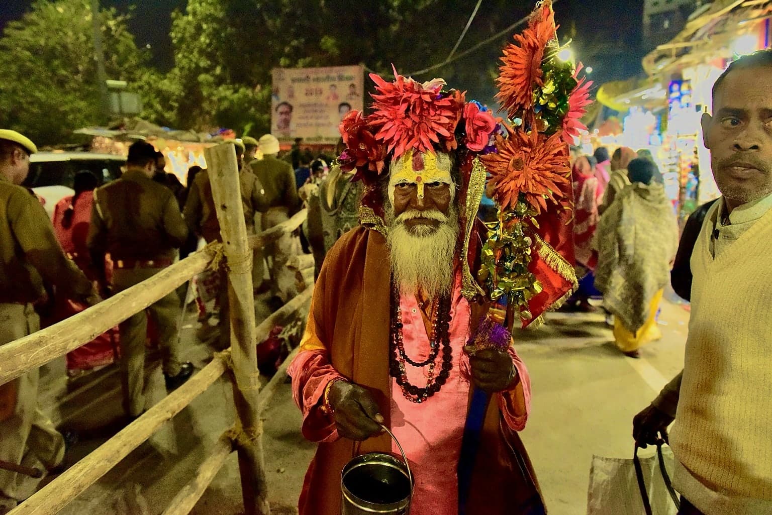 An elderly holy man in vivid clothing and flower decorations faces the camera on a crowded night street in Varanasi, 2019