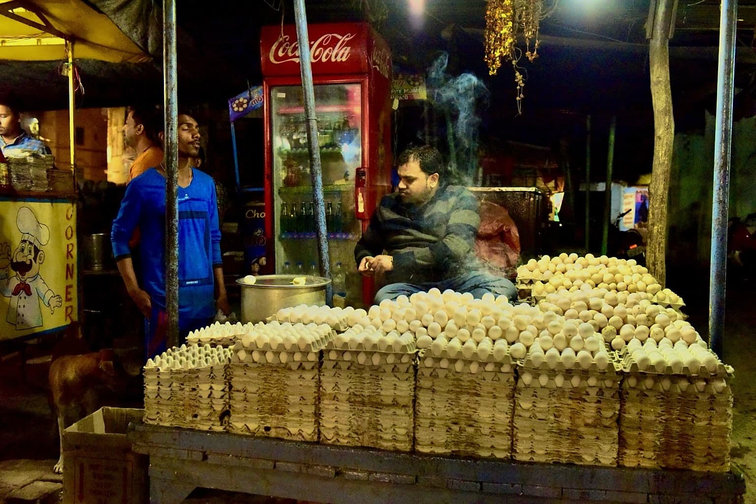 A vendor sits behind a large display of eggs at a brightly lit night stall in Varanasi, 2019