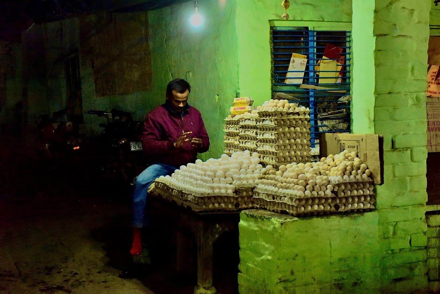 A man sits beside stacks of eggs under a green-lit shelter in Varanasi at night, 2019