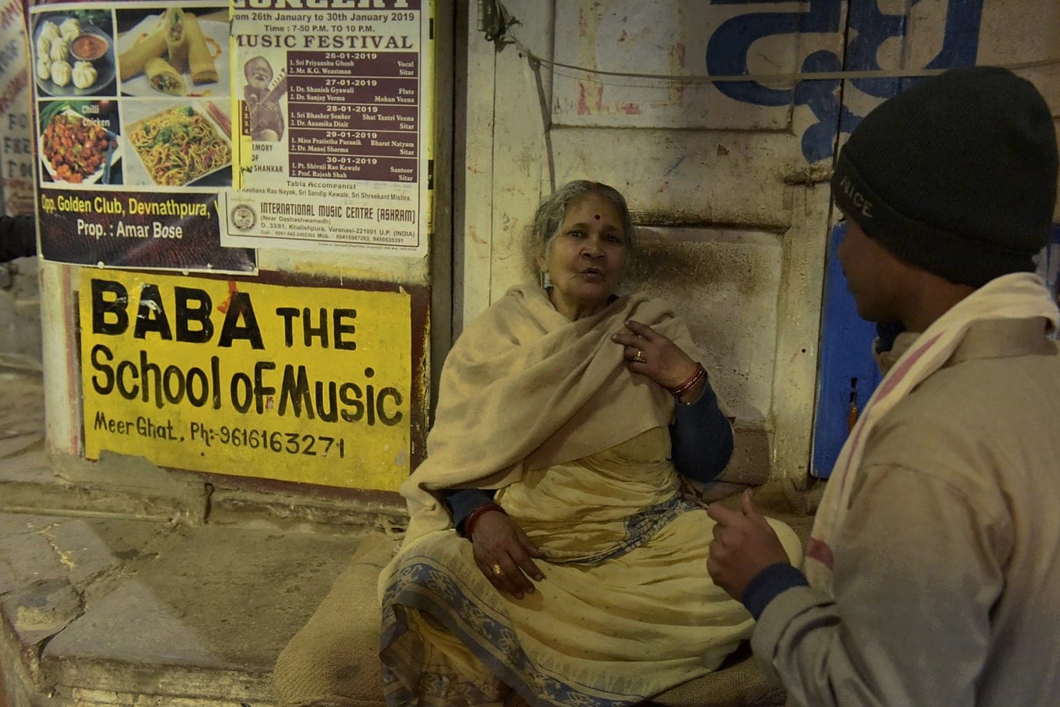 An older woman sits in conversation beside a painted Baba School of Music sign in a Varanasi lane at night, 2019