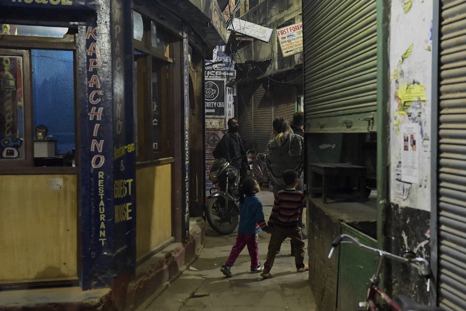 Two children walk through a narrow Varanasi lane lined with small businesses and closed shutters at night, 2019