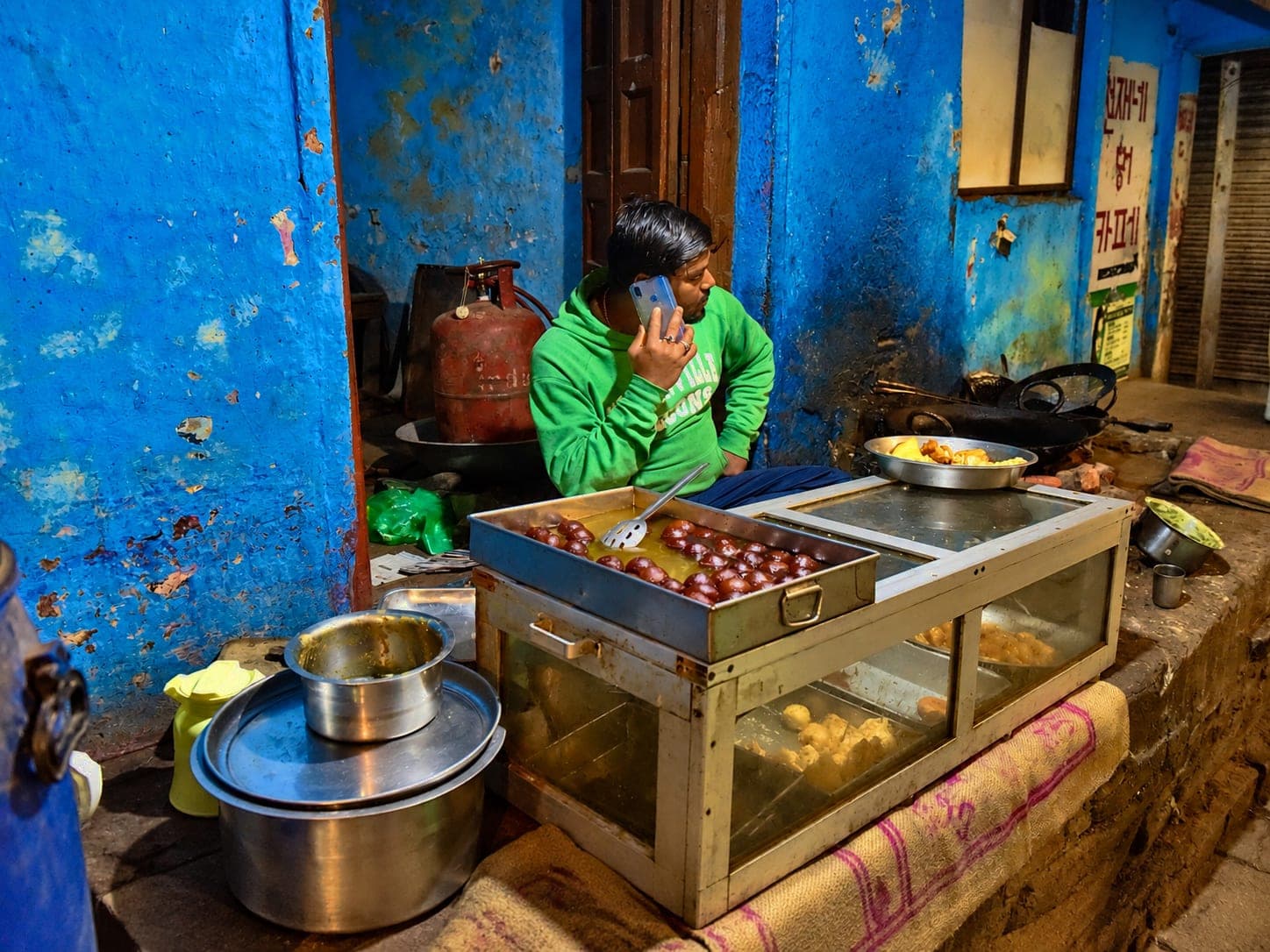 A man in a bright green sweatshirt speaks on a mobile phone beside trays of sweets in a blue-painted night stall in Varanasi, 2019