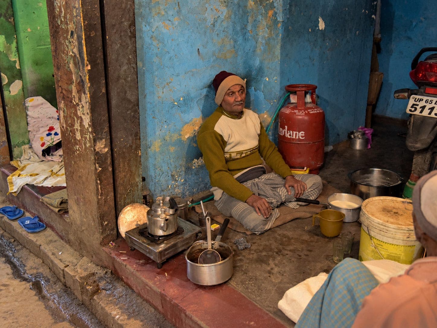 An older man sits in a small cooking space beside metal pots and a red gas cylinder in Varanasi, 2019