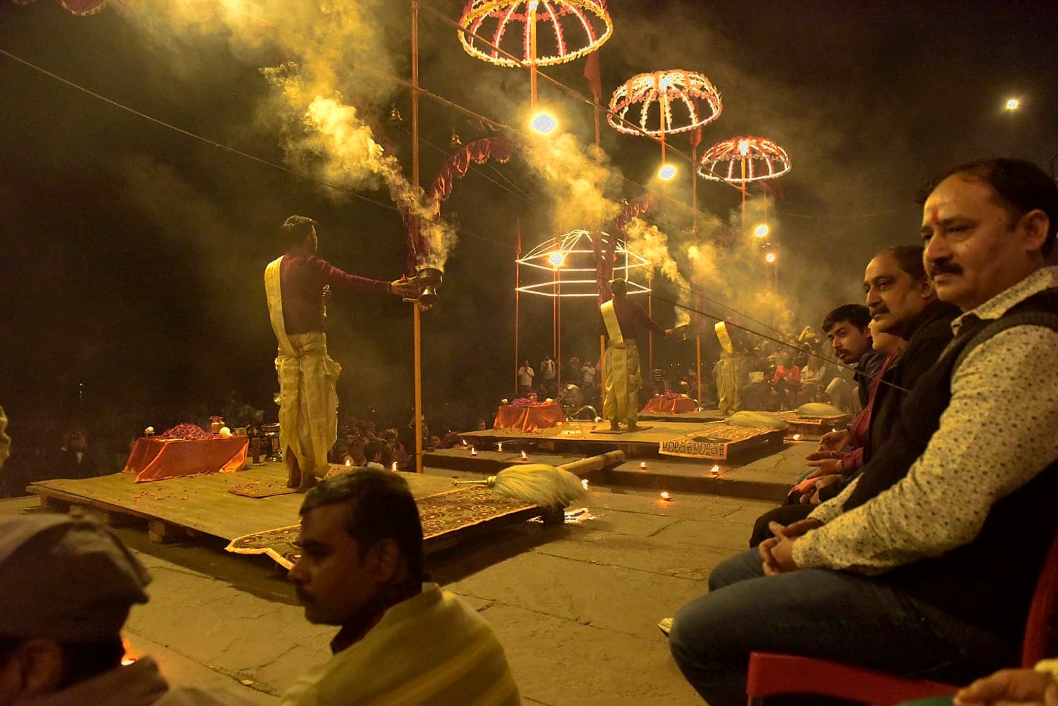 Several priests perform evening Ganga aarti beneath illuminated arches while smoke drifts over the seated crowd in Varanasi, 2019