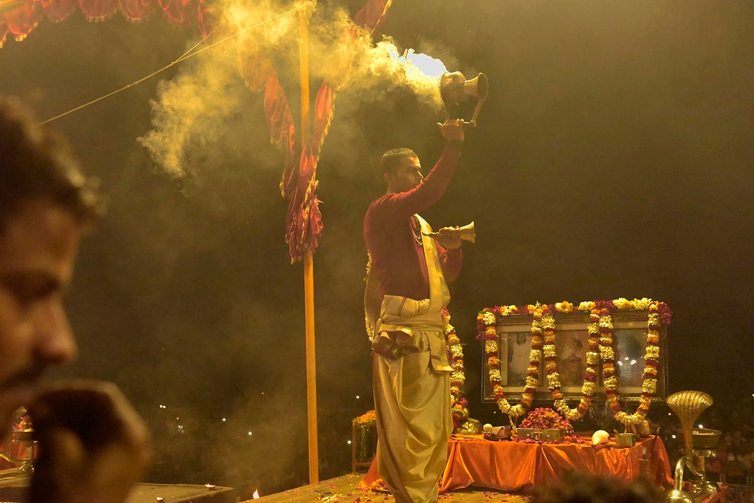 A priest raises a smoking ritual vessel beside a flower-decorated shrine during evening Ganga aarti in Varanasi, 2019