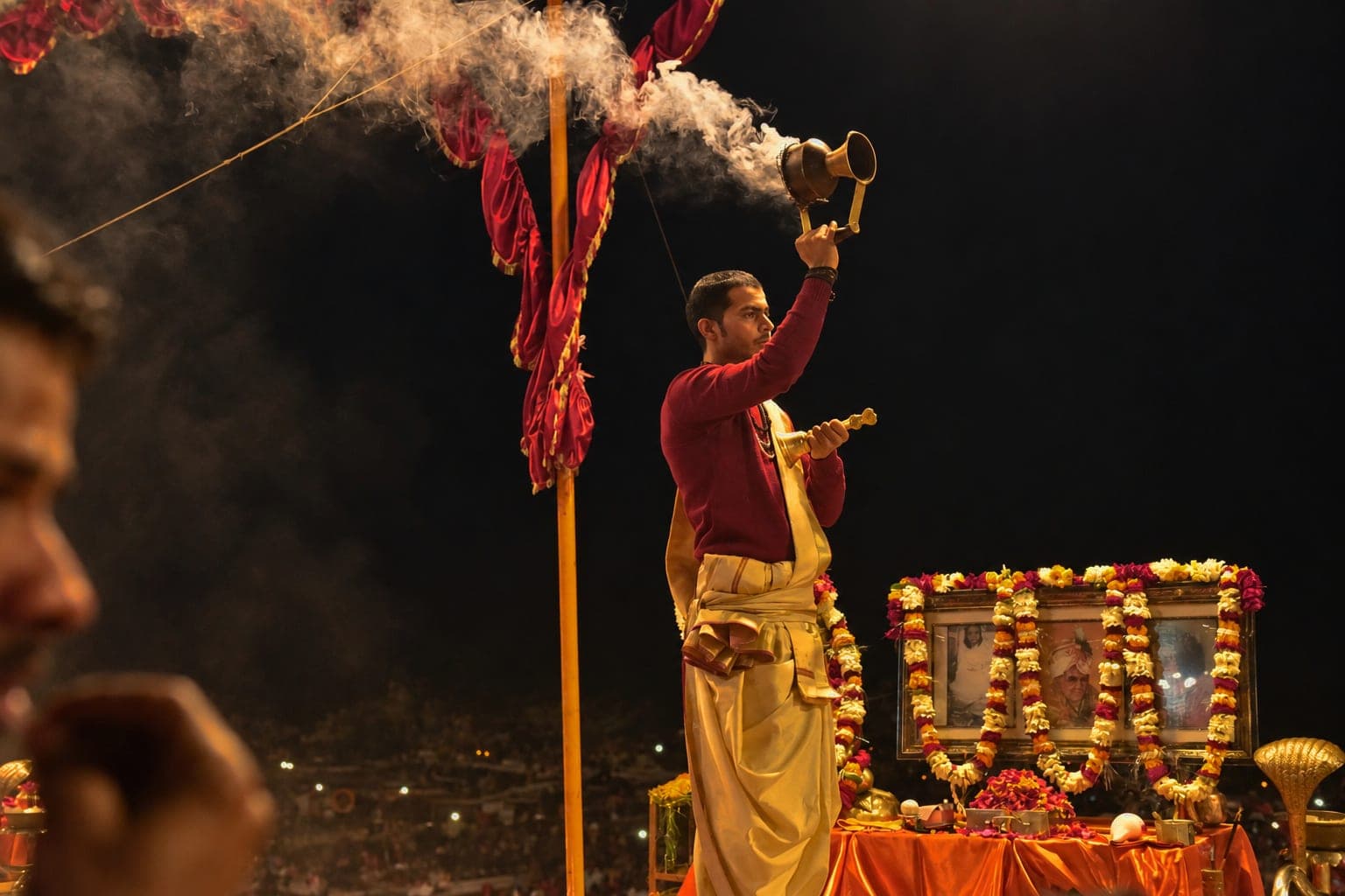 A priest holds a smoking ritual vessel high beside a garlanded shrine during evening Ganga aarti in Varanasi, 2019