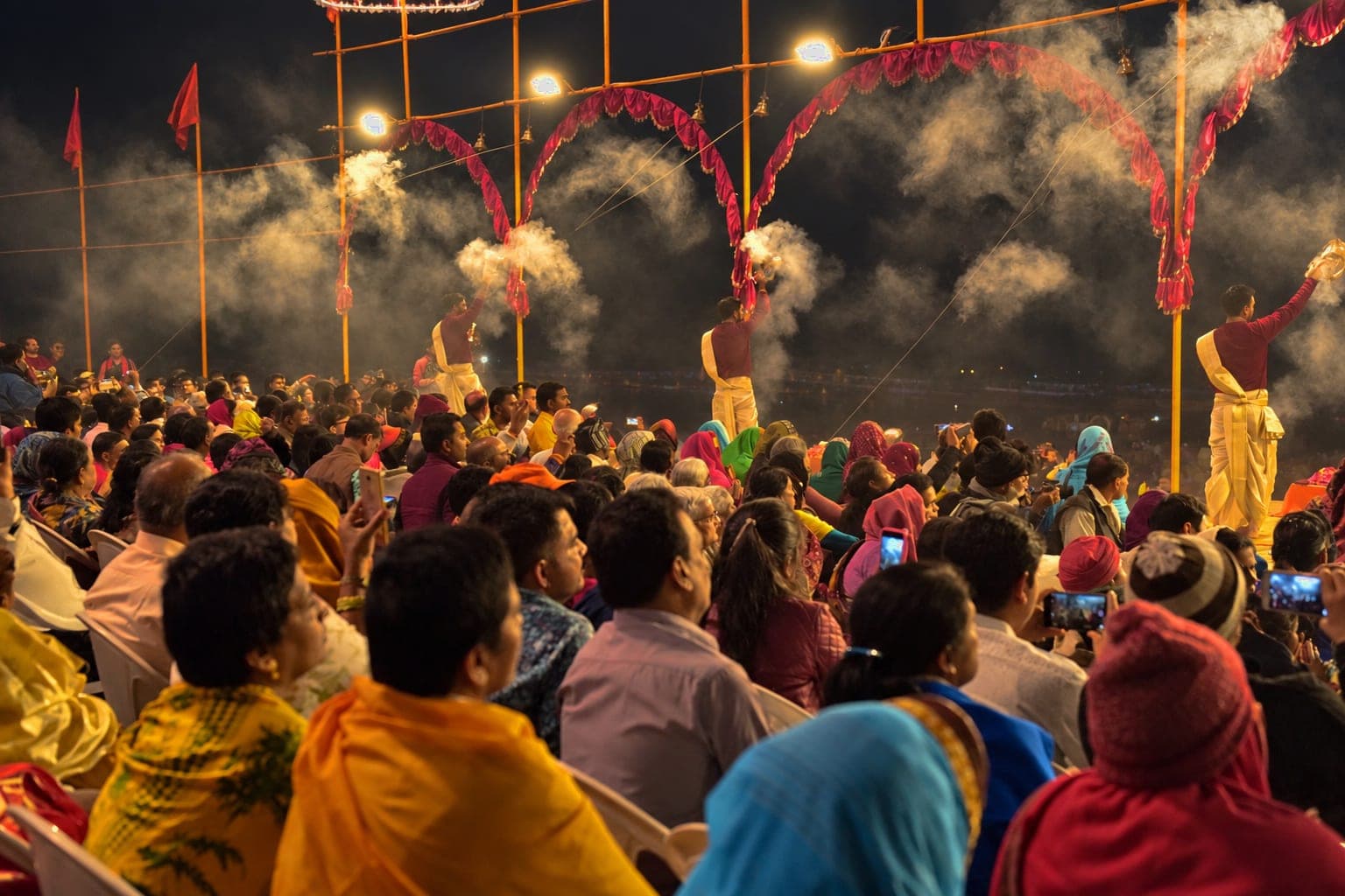 A broad crowd watches several priests as clouds of incense smoke fill the evening Ganga aarti in Varanasi, 2019