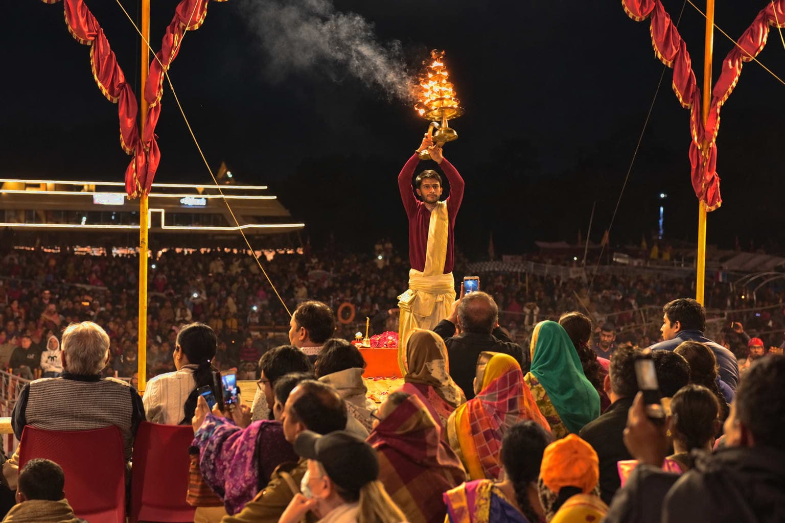 A priest raises a multi-tiered flaming lamp overhead beneath a ceremonial arch during evening Ganga aarti in Varanasi, 2019