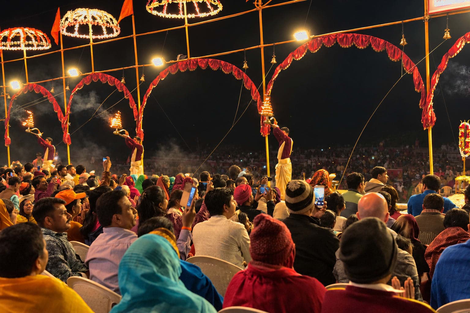 Several priests raise flaming lamps beneath repeating arches as the crowd watches evening Ganga aarti in Varanasi, 2019