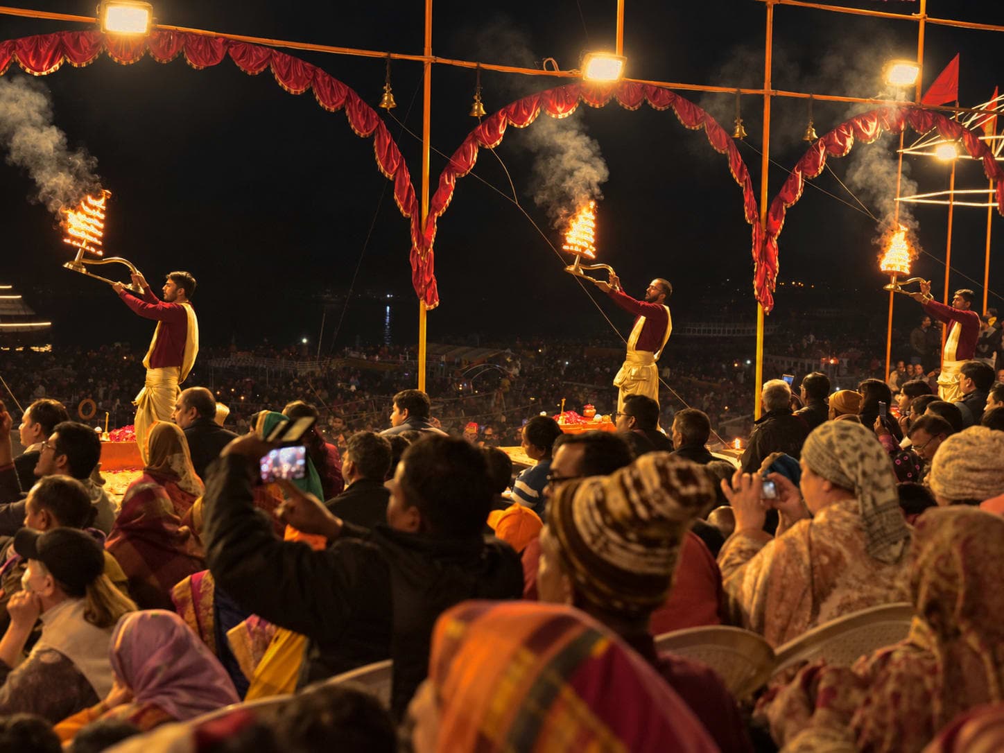 Three priests hold flaming lamps above a dense audience during evening Ganga aarti in Varanasi, 2019