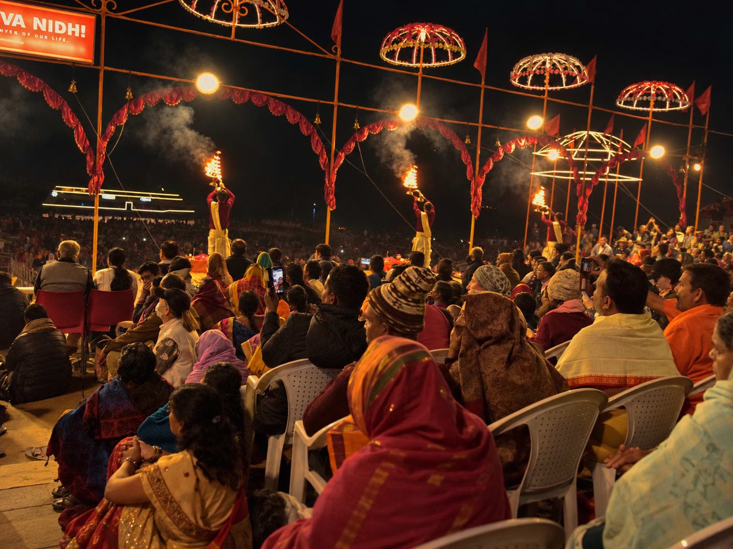 A wide night view shows several priests with flaming lamps beneath illuminated ceremonial structures during Ganga aarti in Varanasi, 2019