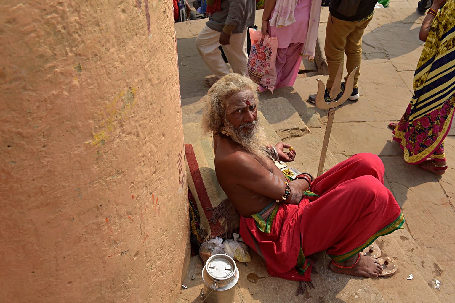 Sadhu seated beside a pillar with his trident, photographed in his surroundings.