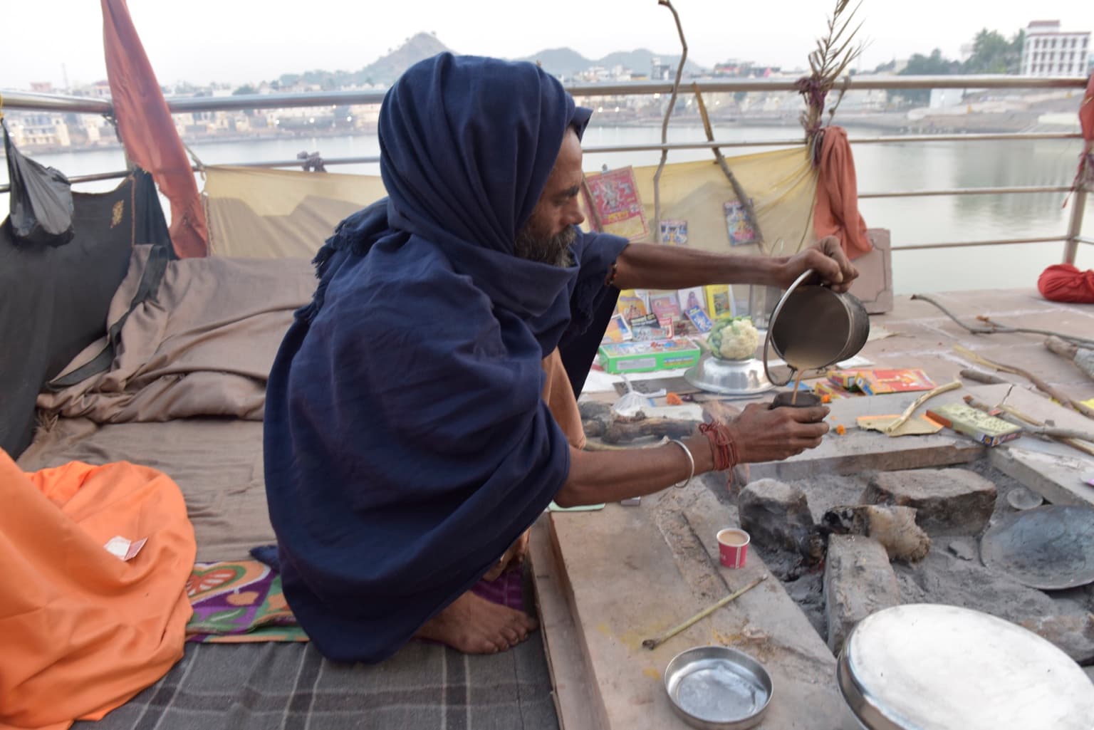 The man in blue pours tea from a metal kettle beside the lake.