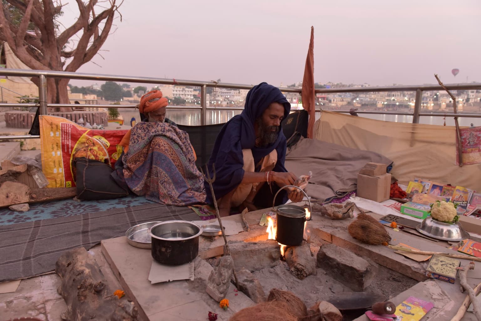 The man in blue tends a pot over a small fire beside the lake while another sadhu sits nearby.