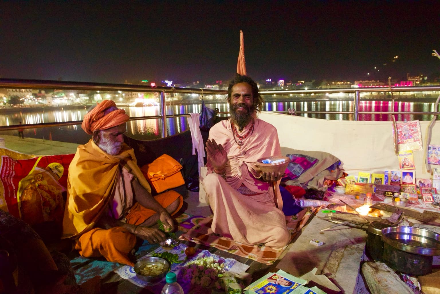 Two sadhus seated beside a fire at night, with Pushkar Lake and its reflected lights behind them.