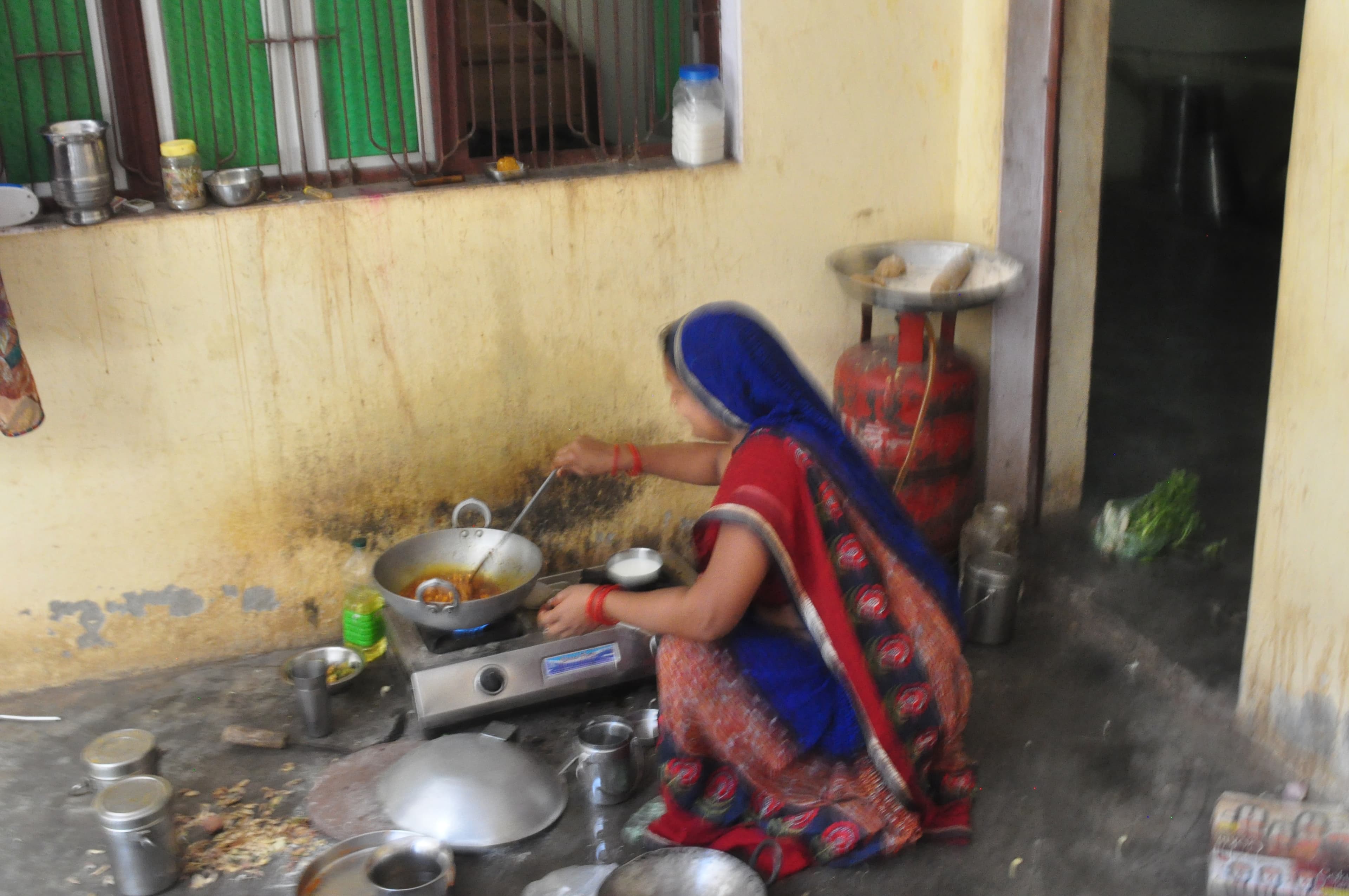 A woman cooking among pots and utensils beside a yellow wall at Baba's home.
