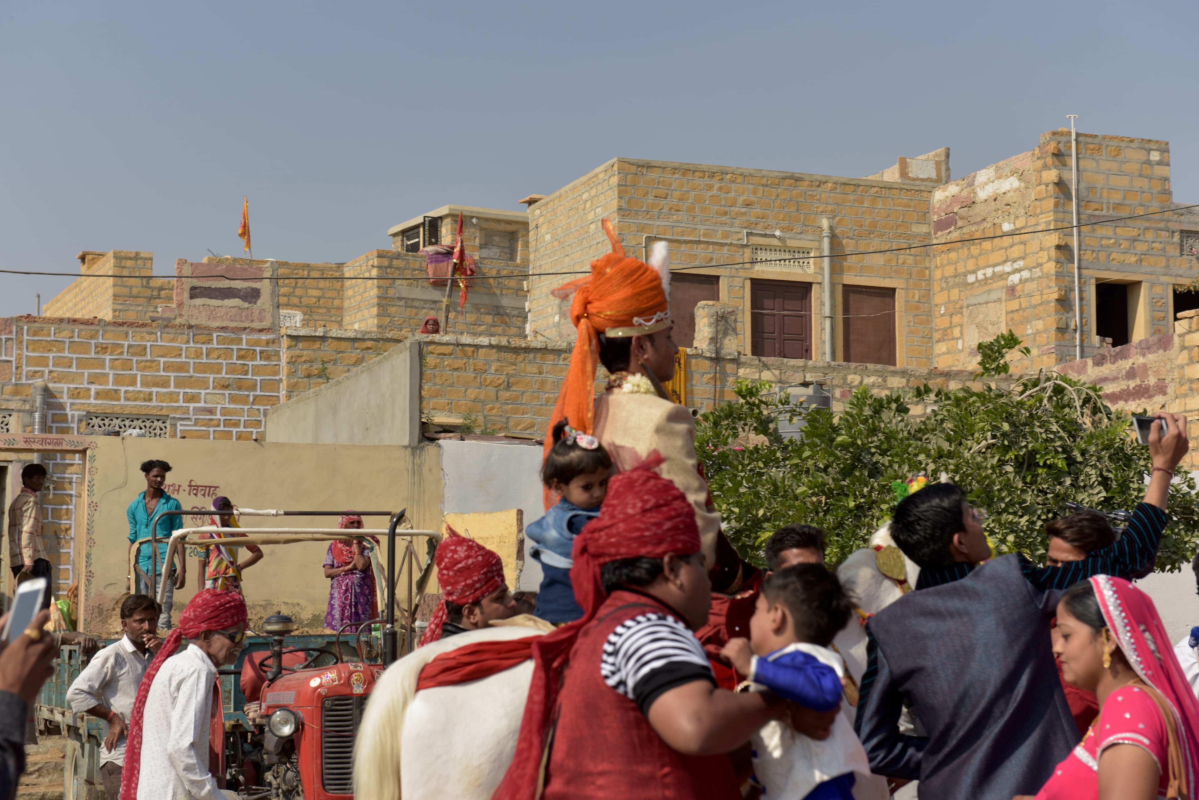 A rider in an orange turban on a white horse, surrounded by people in the wedding procession.