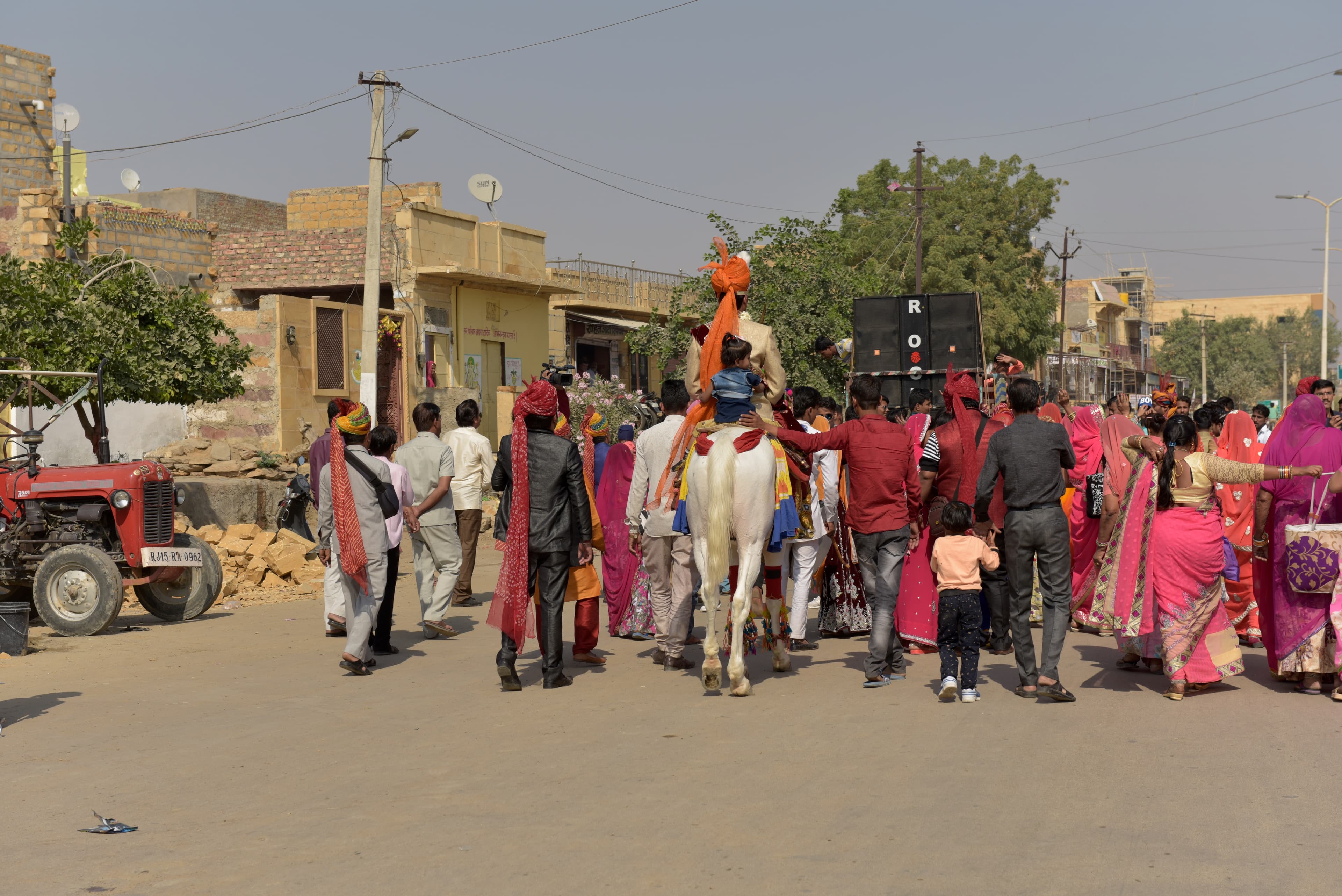 A wedding procession in Jaisalmer, with a rider on a white horse and people walking along the street.