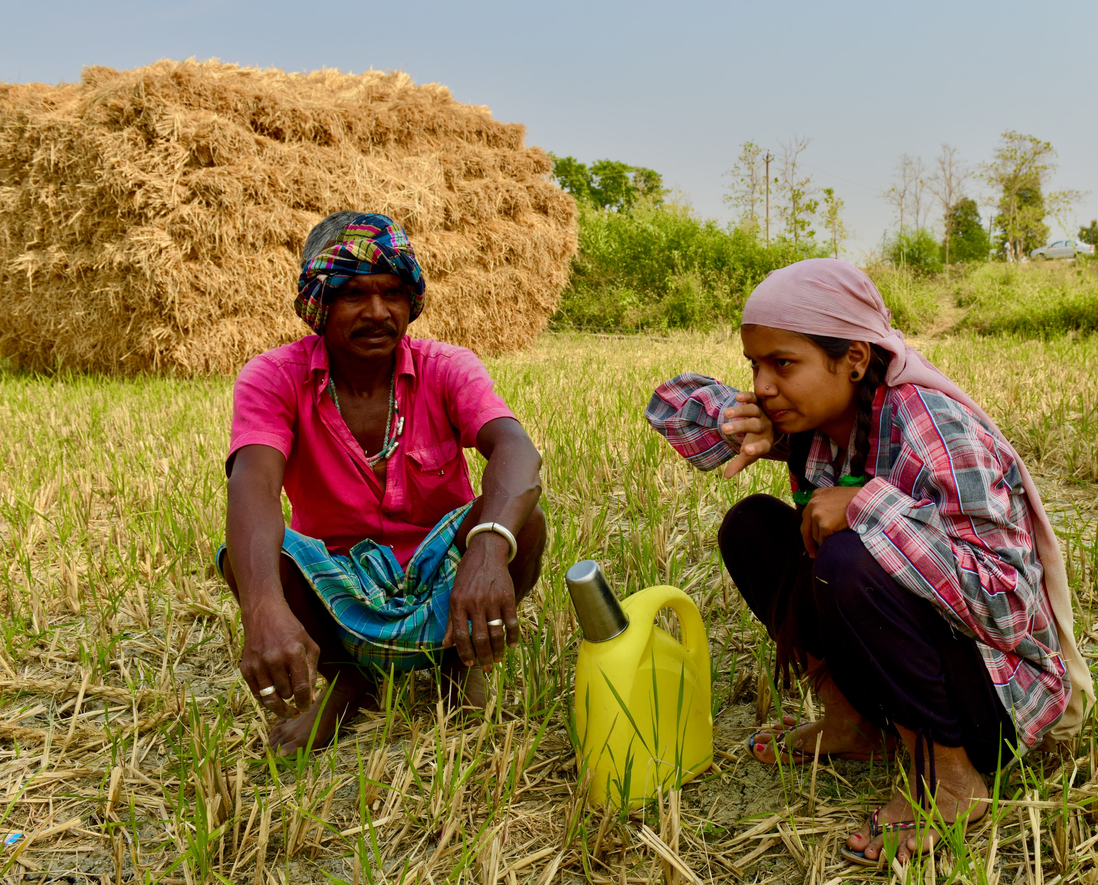 Two agricultural workers resting on a harvested field beside a straw stack and yellow container.
