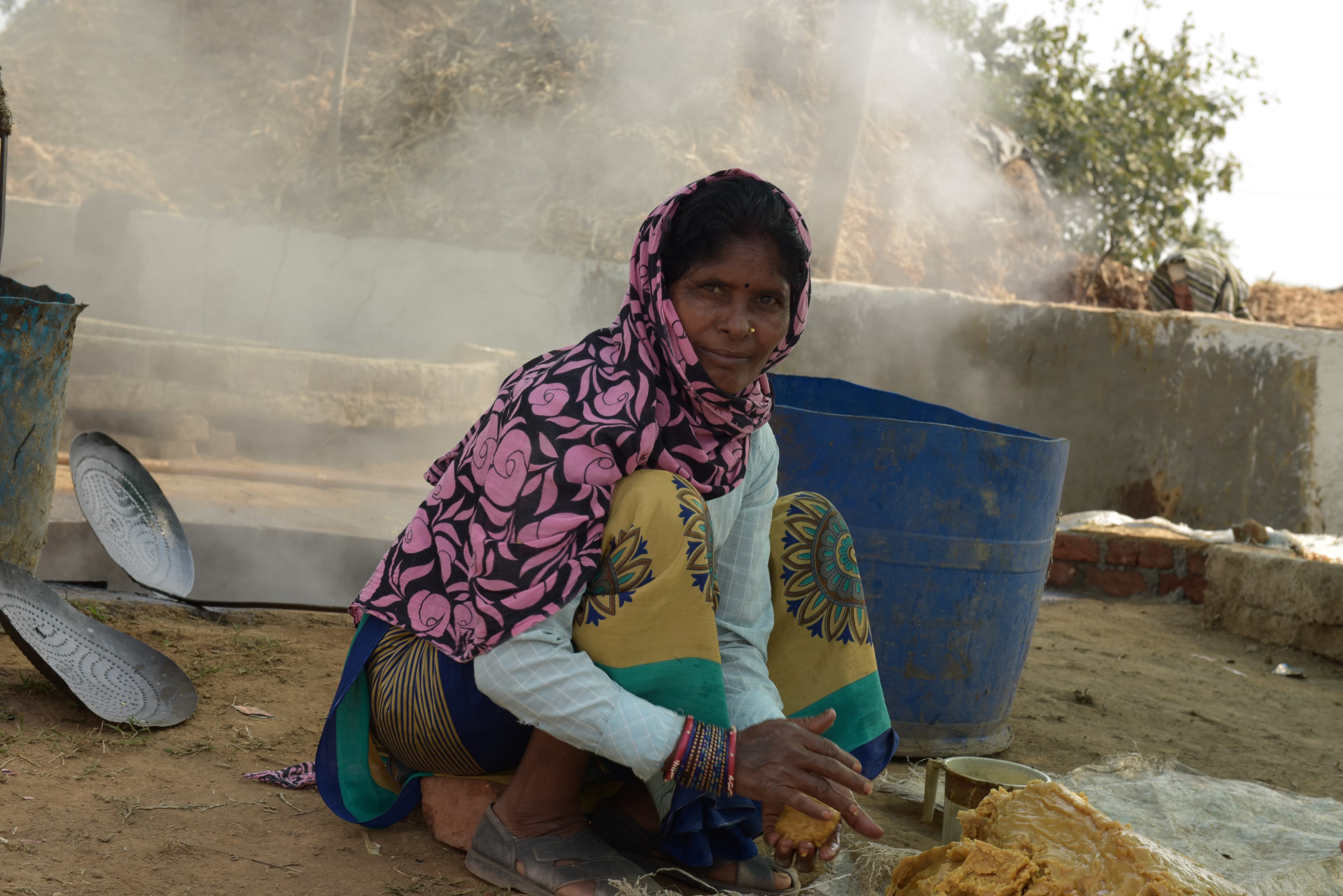 Woman seated by the jaggery preparation area with steam and a blue container behind.