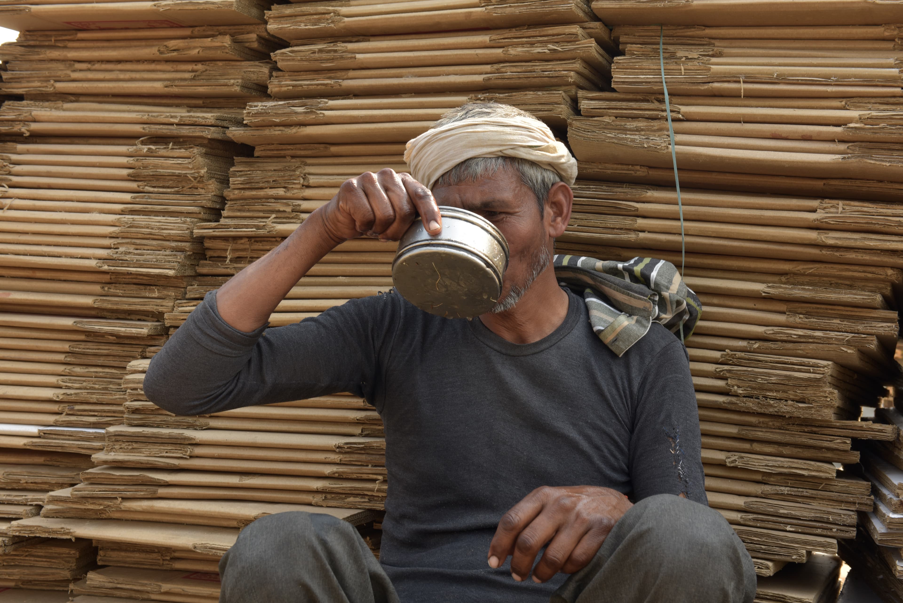 Worker drinking from a metal vessel before stacks of flattened cardboard.